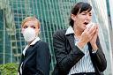 Picture of businesswoman sneezing while another woman wearing a surgical mask looks on