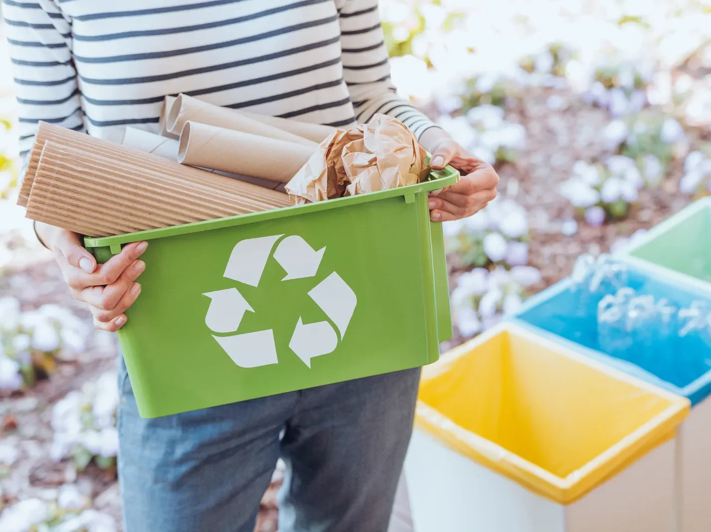 person in a striped shirt and jeans carrying a green recycling container
