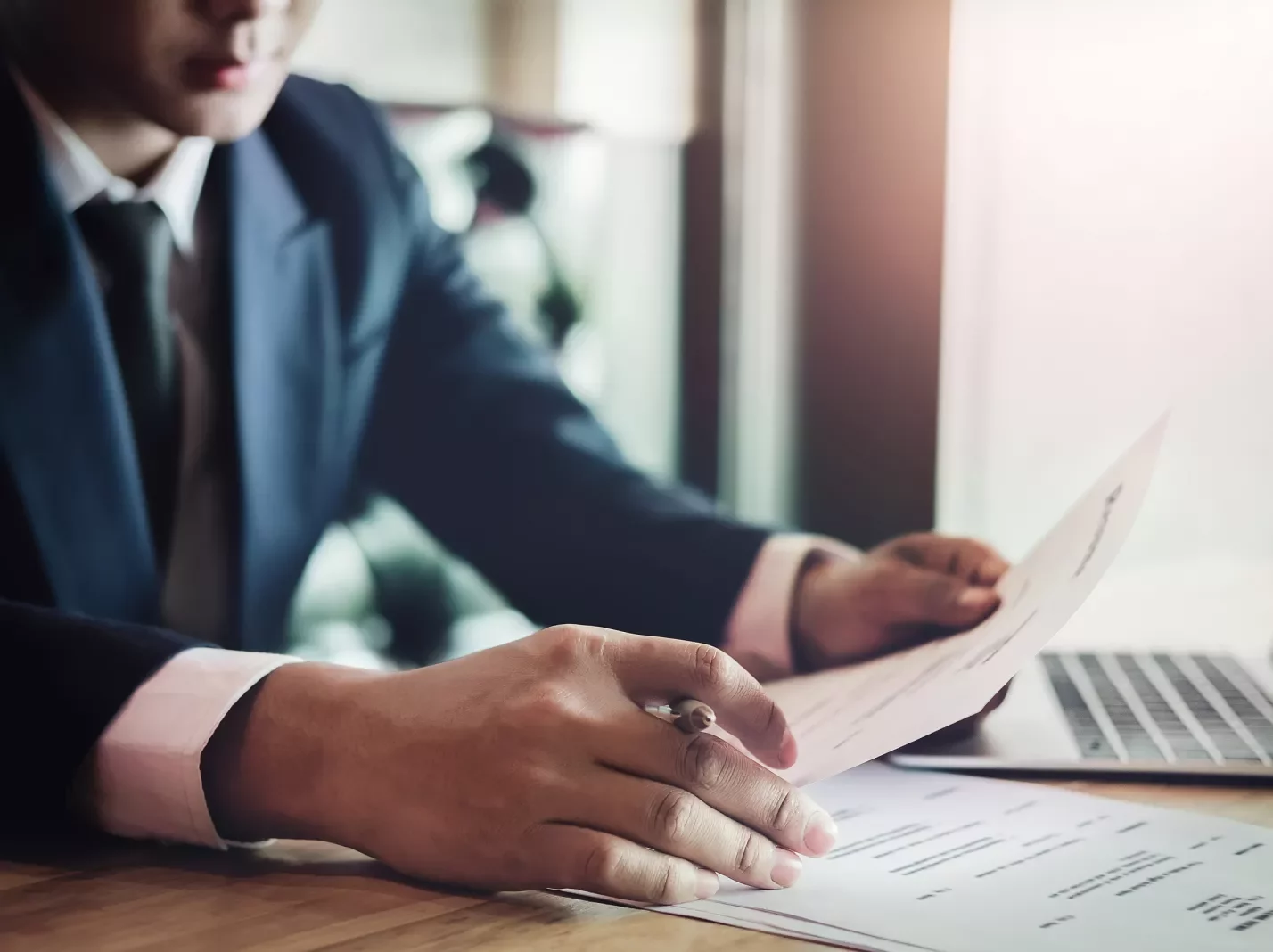 Seated man in a suit holding a pen and documents in front of a laptop