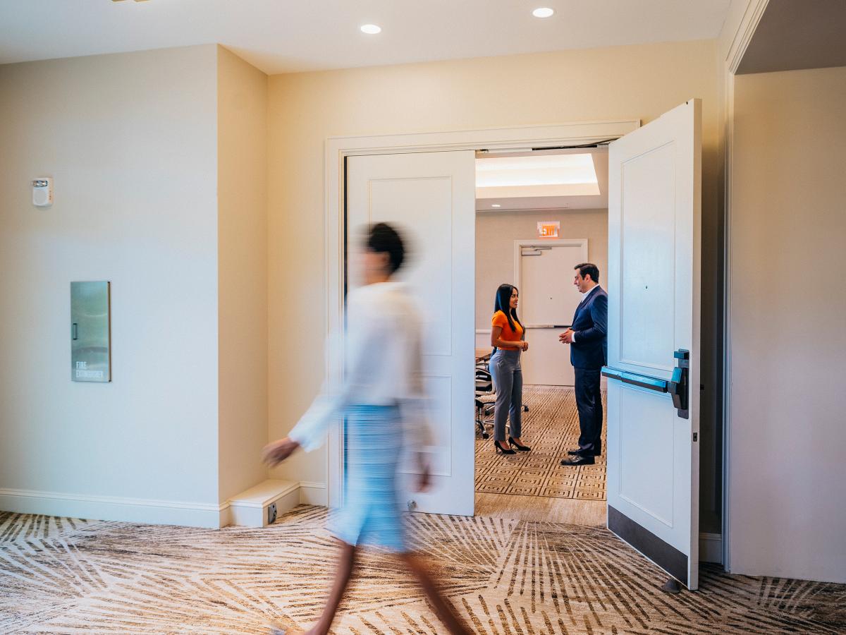 A woman walks past a man and woman talking in an office hallway