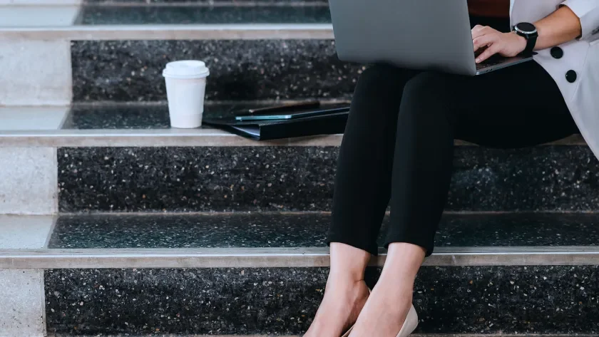 Woman in high heeled shoes sitting on steps next to a cup of coffee with a laptop on her lap