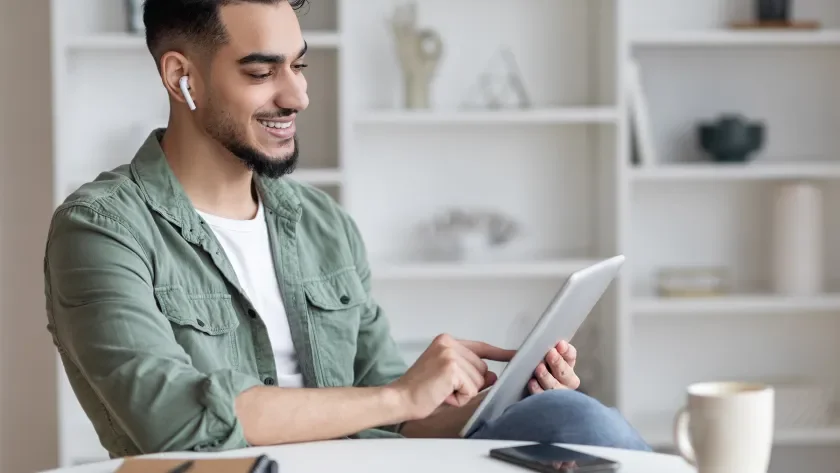 Man in front of a bookcase looking at a tablet and listening on earbuds