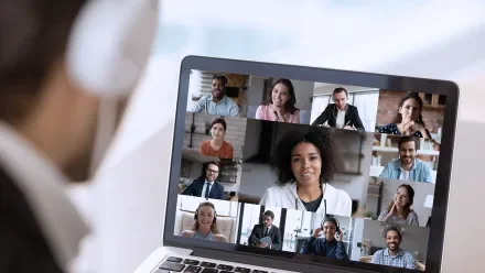 A man in headphones looks at a laptop screen showing a conference call
