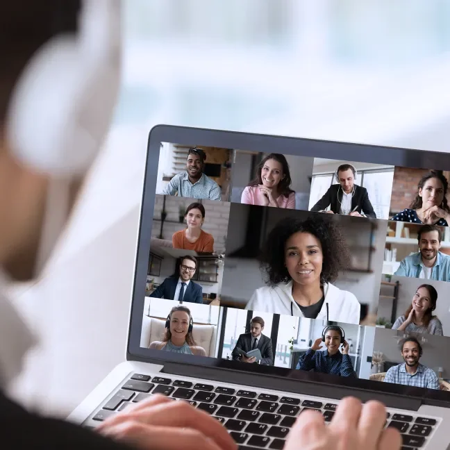 A man in headphones looks at a laptop screen showing a conference call
